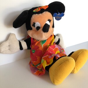 以下が含まれることがあります： カラフルな花柄のドレスと黄色の靴を履いたミニーマウスのぬいぐるみ。おもちゃは白黒のストライプの腕と、耳に付けられたディズニーオリジナルのタグを持っています。