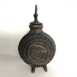 May include: A black and gold metal decorative bottle with intricate floral designs and a small handle on the top. The bottle has three legs and a round body.