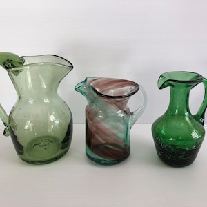 May include: Three green glass jugs with different designs. One jug has a swirl of green and purple glass, another is a solid green, and the third is a clear green with a textured surface.