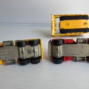 May include: Four toy trucks, two yellow and two red, with black wheels. The trucks are all Hot Wheels brand and have the words "Hot Wheels" and "Made in Malaysia" printed on the bottom.