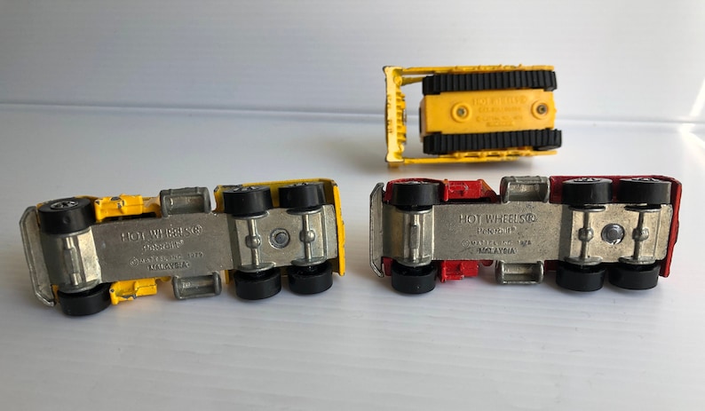 May include: Four toy trucks, two yellow and two red, with black wheels. The trucks are all Hot Wheels brand and have the words "Hot Wheels" and "Made in Malaysia" printed on the bottom.