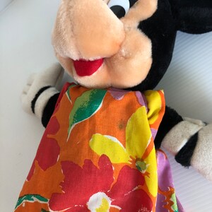 以下が含まれることがあります： カラフルな花柄のドレスを着たミニーマウスのぬいぐるみ。ドレスは明るいオレンジ色の背景に、赤、黄、緑の花が描かれています。