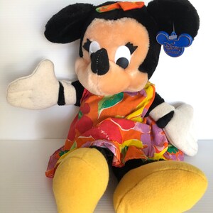 以下が含まれることがあります： カラフルな花柄のドレスと黄色の靴を履いたミニーマウスのぬいぐるみです。おもちゃは黒い鼻、大きな白い目、そして髪に黒いリボンをしています。おもちゃのタグには「ディズニーオリジナル」と書いてあります。