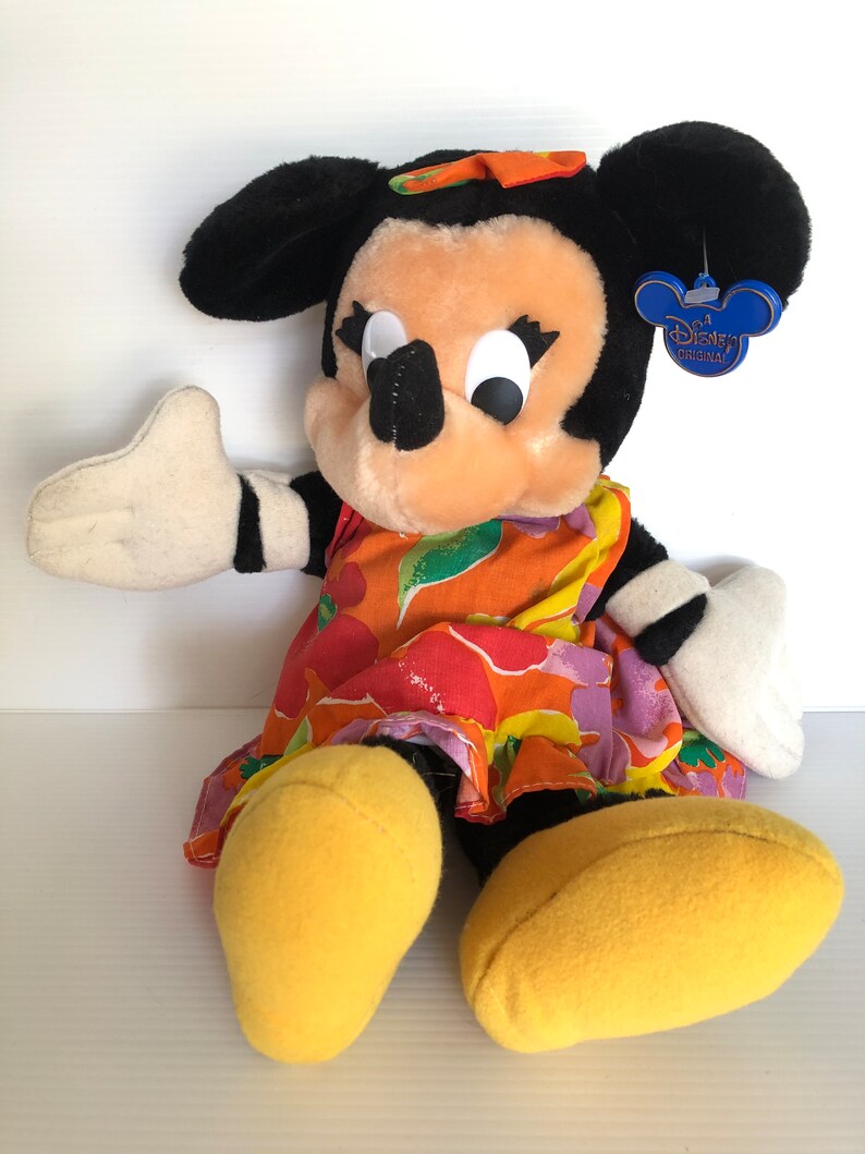 以下が含まれることがあります： カラフルな花柄のドレスと黄色の靴を履いたミニーマウスのぬいぐるみです。おもちゃは黒い鼻、大きな白い目、そして髪に黒いリボンをしています。おもちゃのタグには「ディズニーオリジナル」と書いてあります。
