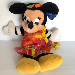 以下が含まれることがあります： 黄色のリボンをつけたカラフルな花柄のドレスを着た、ミニーマウスのぬいぐるみです。おもちゃは白黒のストライプの腕と「ディズニーベビー」と書かれた青いタグが付いています。
