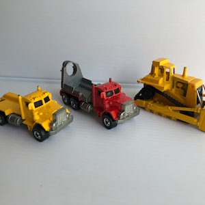 May include: Three toy trucks, a yellow dump truck, a yellow semi-truck with a red cab and a gray flatbed trailer, and a yellow bulldozer.