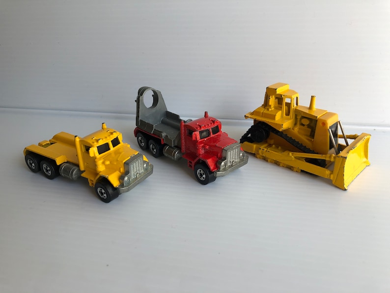 May include: Three toy trucks, a yellow dump truck, a yellow semi-truck with a red cab and a gray flatbed trailer, and a yellow bulldozer.