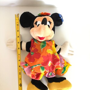 以下が含まれることがあります： 黄色のリボンをつけたカラフルな花柄のドレスを着た、ミニーマウスのぬいぐるみ。人形の高さは約38cmです。