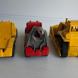 May include: Three toy construction vehicles, a yellow dump truck, a red and gray cement mixer truck, and a yellow bulldozer. The vehicles are all facing forward and are on a white surface.