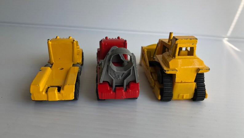 May include: Three toy construction vehicles, a yellow dump truck, a red and gray cement mixer truck, and a yellow bulldozer. The vehicles are all facing forward and are on a white surface.