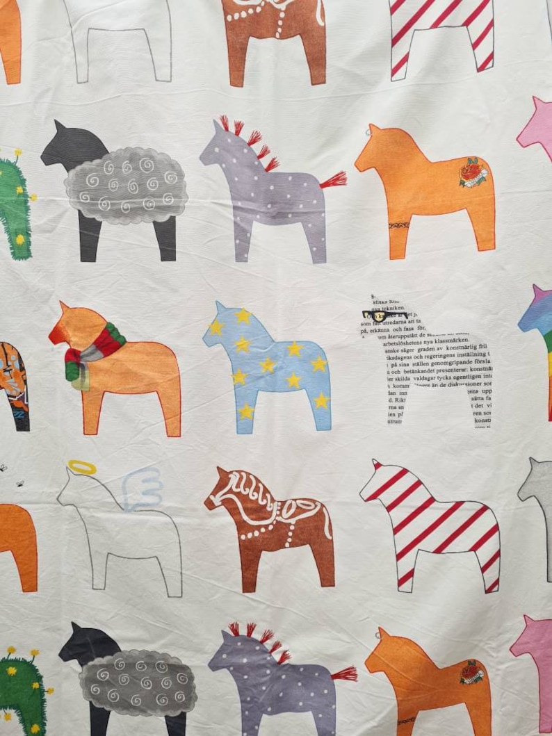 May include: White fabric featuring a repeating pattern of Dala horses in various colours. The horses are decorated with patterns and details, including stars and stripes. The fabric also has some text in the background.