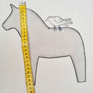 May include: A light grey illustration of a Dala horse with a small bird perched on its back. The horse is approximately 25 cm tall, measured with a yellow measuring tape. The background is white.
