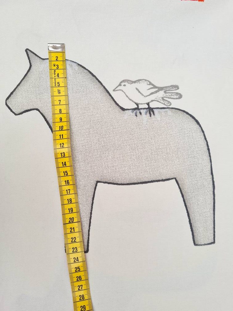 May include: A light grey illustration of a Dala horse with a small bird perched on its back. The horse is approximately 25 cm tall, measured with a yellow measuring tape. The background is white.