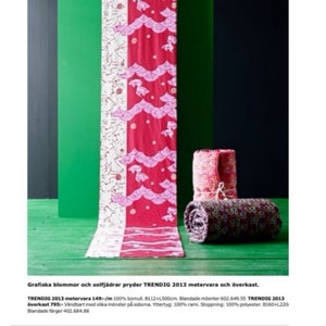 May include: A red and white fabric with a pink and white floral pattern. The fabric is folded in half and has a pink and white floral pattern on one side and a pink and white geometric pattern on the other side. The fabric is on a green surface and there are two rolled up fabric blankets in the background.