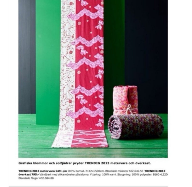 May include: A red and white fabric with a pink and white floral pattern. The fabric is folded in half and has a pink and white floral pattern on one side and a pink and white geometric pattern on the other side. The fabric is on a green surface and there are two rolled up fabric blankets in the background.