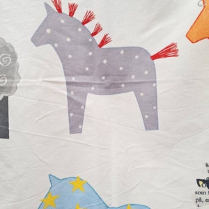 May include: White fabric featuring a repeating pattern of cartoon horses. One horse is grey with white spots and red fringe. Another is blue with yellow star shapes. The design has a playful, childlike aesthetic.
