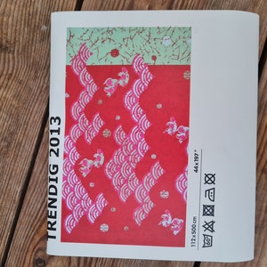 May include: A white brochure with the text "TRENDIG 2013" printed vertically on the left side. The brochure features a red and white patterned fabric with pink fish designs. The fabric measures 44 x 197 inches and 112 x 500 cm. The brochure also includes care instructions for the fabric.