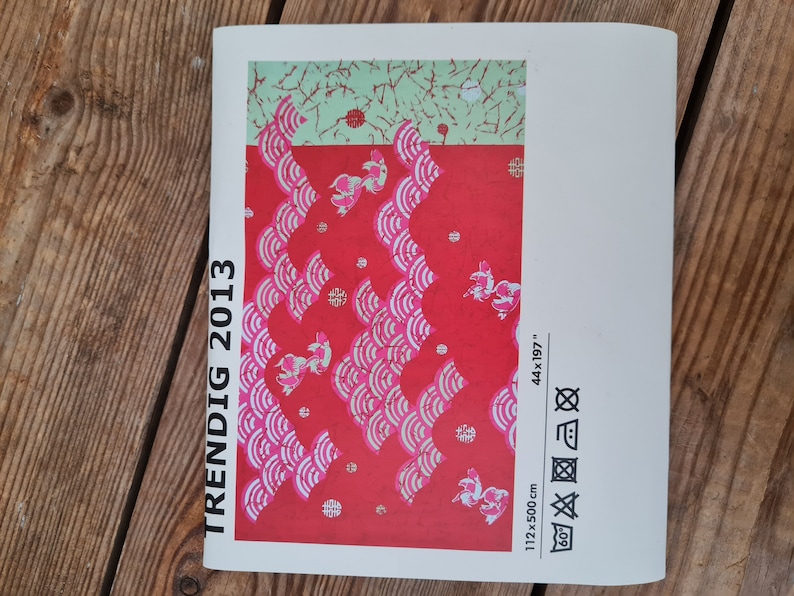 May include: A white brochure with the text "TRENDIG 2013" printed vertically on the left side. The brochure features a red and white patterned fabric with pink fish designs. The fabric measures 44 x 197 inches and 112 x 500 cm. The brochure also includes care instructions for the fabric.