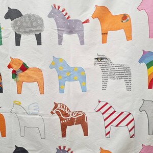 May include: White fabric with a repeating pattern of colourful, stylised Dala horses. The horses are in various colours, including orange, blue, and pink, with decorative details. Some text is visible in the background.