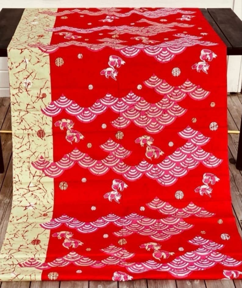 May include: Red fabric with a repeating pattern of pink and white waves and a stylized fish design. The fabric has a gold and white border.
