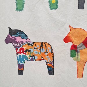 May include: White fabric featuring a repeating pattern of Dala horses. One horse is adorned with graffiti art in purple, orange, and blue. Other horses are in solid colours, including green and orange, with simple designs.