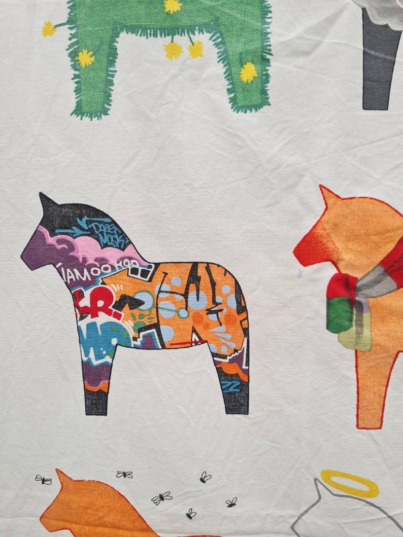 May include: White fabric featuring a repeating pattern of Dala horses. One horse is adorned with graffiti art in purple, orange, and blue. Other horses are in solid colours, including green and orange, with simple designs.