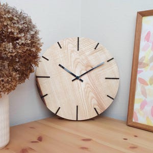 Könnte beinhalten: Eine runde Holzuhr mit schwarzen Zeigern und schwarzen Stundenmarkierungen. Die Uhr ist aus hellem Holz mit einer natürlichen Maserung. Die Uhr hängt an einer Wand.