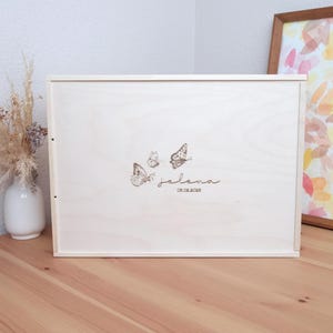 Peut inclure: Une boîte en bois blanche avec un motif de papillon et le nom "Jelena" gravé sur le devant. La boîte est fermée et posée sur une surface en bois.