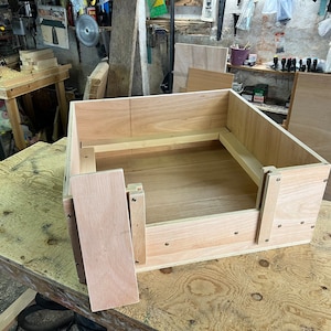 May include: A partially constructed wooden box, possibly for storage or a small pet bed, is positioned on a wooden workbench. The box is made from light-coloured wood, with visible joints and screws. A detached side panel rests close by, indicating ongoing construction.