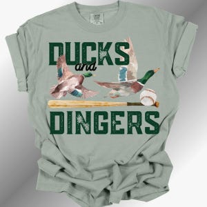 May include: A sage green short-sleeve t-shirt featuring a graphic of two mallard ducks in flight, a wooden baseball bat, and a baseball. The text "DUCKS and DINGERS" is printed in dark green block letters above and below the illustrated elements.