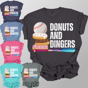 May include: A collection of t-shirts in various colors, each featuring a baseball, donuts, and a baseball bat graphic. The text "DONUTS AND DINGERS" is printed on each shirt. The shirts are in navy, blue, pink, teal, and gray.