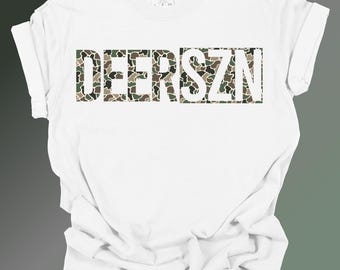 May include: A white short-sleeve t-shirt featuring the text DEERSZN printed in a camouflage pattern of green, brown, and black. The shirt has a crew neckline and a standard fit, with the brand label visible on the inside of the collar area.
