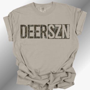 Könnte beinhalten: Ein hellbeiges T-Shirt mit kurzen Ärmeln und dem Text DEERSZN in Blockschrift. Die Buchstaben sind mit einem braunen, grünen und beigen Tarnmuster gefüllt. Das T-Shirt hat einen Rundhalsausschnitt und eine Standardpassform mit hochgekrempelten Ärmeln.
