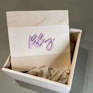 Puede incluir: Una caja de madera con tapa, la tapa tiene el nombre "Riley" escrito en letras moradas. La caja está abierta y tiene un tejido beige en su interior.