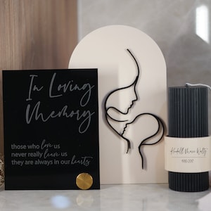 May include: A black acrylic plaque with the text "In Loving Memory" and the quote "those who love us never really leave us they are always in our hearts". A white wooden cutout of a mother and child in a line art style. A black pillar candle with the text "Koufalll Thom Walty 1980-2017" on a white label.