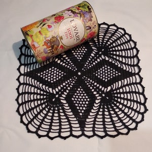 Può includere: Tovaglietta all'uncinetto nera con un design delicato e intricato. La tovaglietta è circolare con un foro centrale e un bordo a festoni. È posizionata piatta su una superficie bianca.