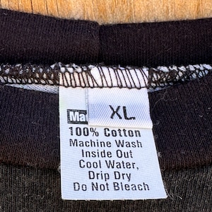 May include: Close-up of a black t-shirt collar with a white tag. The tag reads "Ma XL 100% Cotton Machine Wash Inside Out Cool Water, Drip Dry Do Not Bleach". The shirt is made of a dark fabric.