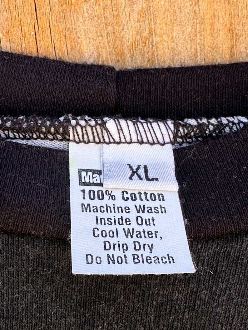 May include: Close-up of a black t-shirt collar with a white tag. The tag reads "Ma XL 100% Cotton Machine Wash Inside Out Cool Water, Drip Dry Do Not Bleach". The shirt is made of a dark fabric.