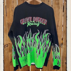 May include: Black long-sleeve shirt featuring a green flame design and the text "Grave Digger Monster Truck Racing." The shirt has signatures and the text "Grave Digger Exclusive" at the top.