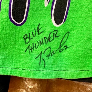 May include: Close-up of a neon green t-shirt with the words "BLUE THUNDER" and a signature in black ink. The shirt has a black and white design with purple accents.