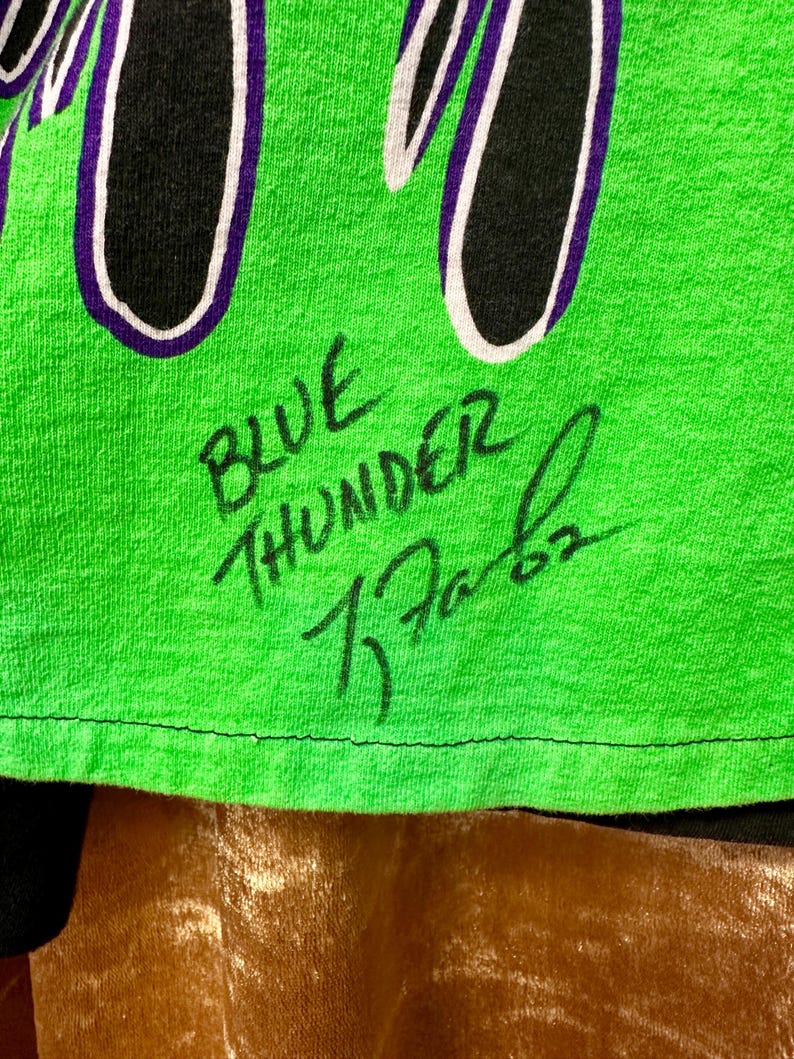 May include: Close-up of a neon green t-shirt with the words "BLUE THUNDER" and a signature in black ink. The shirt has a black and white design with purple accents.