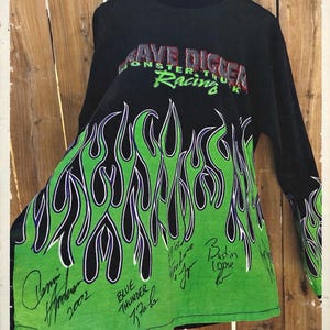 May include: Black long-sleeve shirt with a green and black flame design. The shirt features the text "Grave Digger Monster Truck Racing" in red and green. Several signatures are visible on the green portion of the shirt.