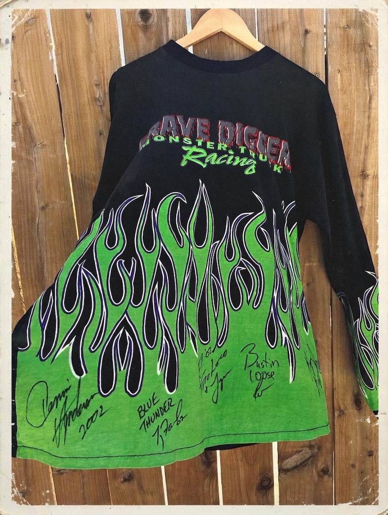 May include: Black long-sleeve shirt with a green and black flame design. The shirt features the text "Grave Digger Monster Truck Racing" in red and green. Several signatures are visible on the green portion of the shirt.