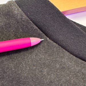 May include: Close-up of a dark grey t-shirt with a black collar. A bright pink pen is in the foreground, pointing towards the shirt. The fabric has a textured appearance.