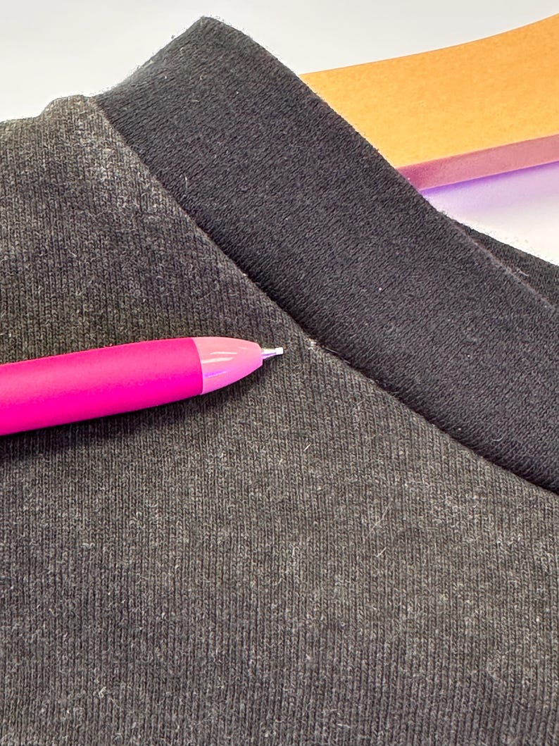 May include: Close-up of a dark grey t-shirt with a black collar. A bright pink pen is in the foreground, pointing towards the shirt. The fabric has a textured appearance.
