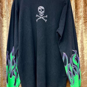 May include: Black long-sleeved shirt with green and purple flame design on the sleeves. The back of the shirt features a skull and crossbones graphic with an eye patch.