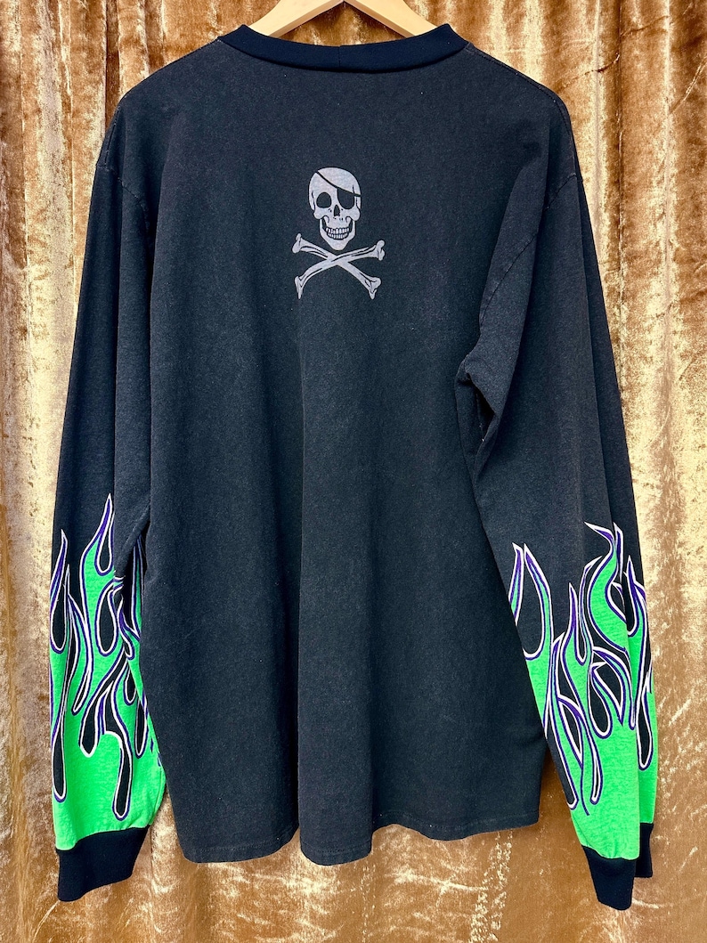 May include: Black long-sleeved shirt with green and purple flame design on the sleeves. The back of the shirt features a skull and crossbones graphic with an eye patch.