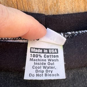May include: A white clothing label with black text, attached to a black garment. The label states "Made in USA", "100% Cotton", and washing instructions. The garment is black.