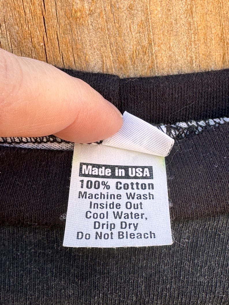 May include: A white clothing label with black text, attached to a black garment. The label states "Made in USA", "100% Cotton", and washing instructions. The garment is black.
