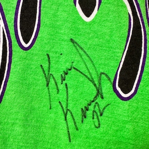 May include: Close-up of a vibrant green t-shirt with a flame design in black, purple, and white. The shirt has a handwritten signature in black ink.